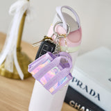Purple holographic keychain attached to a white shoe with a blurred Prada bag in the background.
#color_iridescent