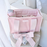 A light pink fabric organizer tote bag with satin ribbon handles and a bow, shown open with storage compartments and accessories inside, placed on a round stool in a bright indoor setting.
#color_ballet pink bow