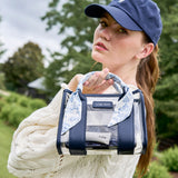 A woman outdoors wearing a navy baseball cap and cream cable-knit sweater, holding a transparent handbag with navy trim and a blue-and-white patterned scarf. The bag has a visible CASE+MATE logo patch.
#color_something navy twilly