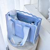 A light blue tote bag with white handles and a white zipper, shown filled with travel accessories like a water bottle, earbuds case, and small toiletries. The bag is styled on a round stool in a bright, modern room.
#color_carolina blue