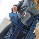 A person wearing a gray sweater holds a large denim-style tote bag with clear side pockets and a blue zippered pouch inside. The photo is cropped to show the person's torso and hands in a bright indoor setting.
#color_oxford navy