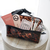 A clear cosmetic pouch with a brown leopard print pattern and black trim, shown open on a round stone pedestal. Inside are makeup brushes, a pink tube labeled 'SUMMER FRIDAYS,' a striped card, and a small clear bag.
#color_tortoiseshell