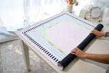 A person unrolls a large rectangular mat on a white table in a bright indoor setting. The mat has a pastel pink and light blue striped center, a navy blue border with white dots, and a pale green curved accent section.
#color_classic cabana stripe