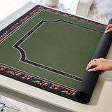 A green felt playing card mat with a black and orange patterned border, partially unrolled on a light-colored table. A person's hands are visible holding the rolled edge of the mat.
#color_classic tortoiseshell