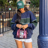 A woman wearing a navy and green color-block sweatshirt, a green 'Ralph Lauren' cap, and holding a burgundy patterned clear stadium bag, standing outdoors near a blue column.
#color_mulberry twilly