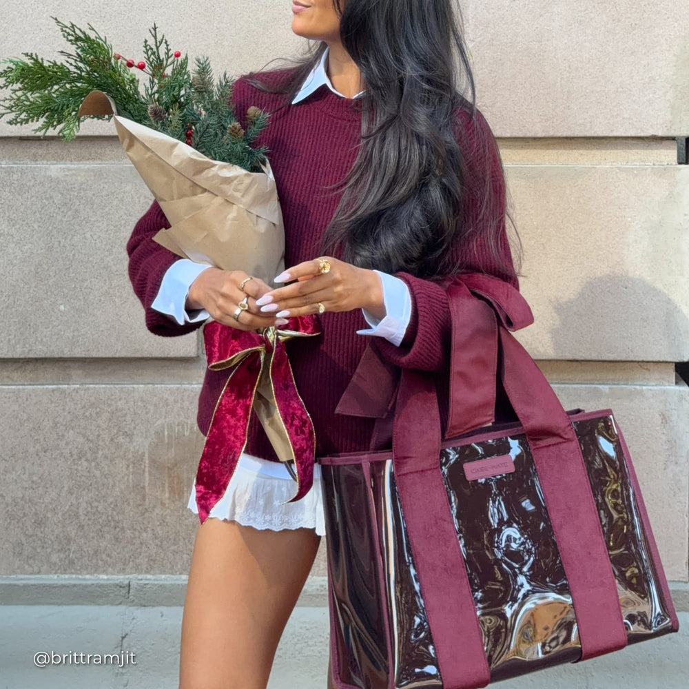 A woman wearing a burgundy ribbed sweater and white collared shirt holds a wrapped holiday bouquet and carries a large burgundy and glossy clear tote bag, standing against a beige stone wall.
#color_holiday edition red velvet bow