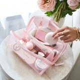 A pink translucent makeup bag filled with cosmetics, placed on a round marble table. A person's hand is holding one of the cosmetic items inside the bag.
#color_ballet pink bow