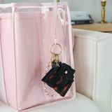 A pink translucent zippered tote bag with a black and brown tortoiseshell miniature suitcase keychain charm attached to the key leash, resting on a light beige sofa
#color_tortoiseshell bow