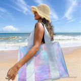 A woman on a sunny beach wearing a straw sun hat and carrying a large iridescent holographic tote bag with white straps printed with the brand name CASE MATE
#color_carolina blue iridescent