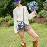 A person outdoors on grass wearing a cream cable-knit sweater, brown gingham shorts, brown cowboy boots, a navy cap, and a blue crossbody bag, while holding a blue and white pom-pom.
#color_something navy twilly