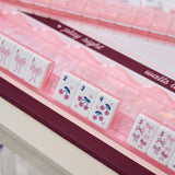 Pink and white mahjong tiles with cherry and bow designs, arranged in a clear plastic case with a dark burgundy zipper and decorative ribbon.
#color_ballet pink pearl edition