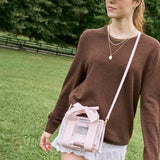A woman wearing a brown knit sweater and white skirt carries a small transparent pink crossbody bag with a bow on a strap in an outdoor park setting.
#color_ballet pink bow