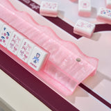 A pink translucent acrylic mahjong rack holding several white and pink mahjong tiles on a light tabletop with a dark burgundy runner underneath.
#color_Ballet Pink Pearl