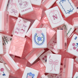 A close-up pile of pink and white acrylic mahjong tiles with decorative symbols and illustrations, including bows, cherries, dragons, and floral motifs.
#color_ballet pink pearl edition
