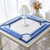 A square white table mat with blue decorative corner sections and a central blue border, displayed on a table in a bright room with a small white lamp and a vase of flowers.
#color_classic carolina blue