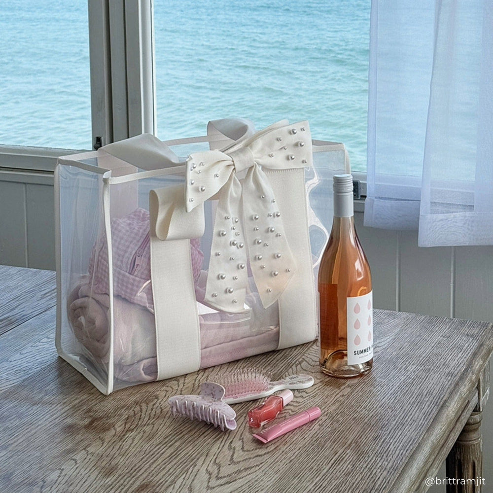 Gift bag with a bow, bottle of rose, and beauty items on a table by a window with water view. #color_ivory pearl bow