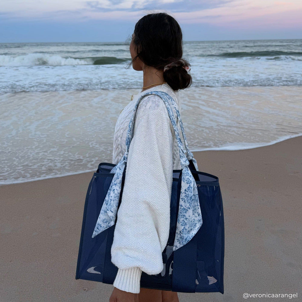Person standing on a beach with a blue tote bag and floral towel, facing the ocean.
#color_something navy twilly