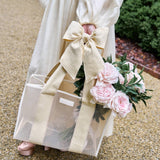 Person holding a gift bag with a large bow and pink flowers, standing on a gravel path.
#color_raffia bow