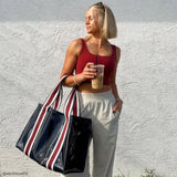 Woman in red top and white pants holding a striped bag and a drink against a light-colored wall.
#color_navy rugby stripe