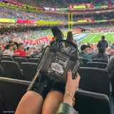 Person holding a black handbag with a clear panel at a sports event in a stadium. #color_midnight_black