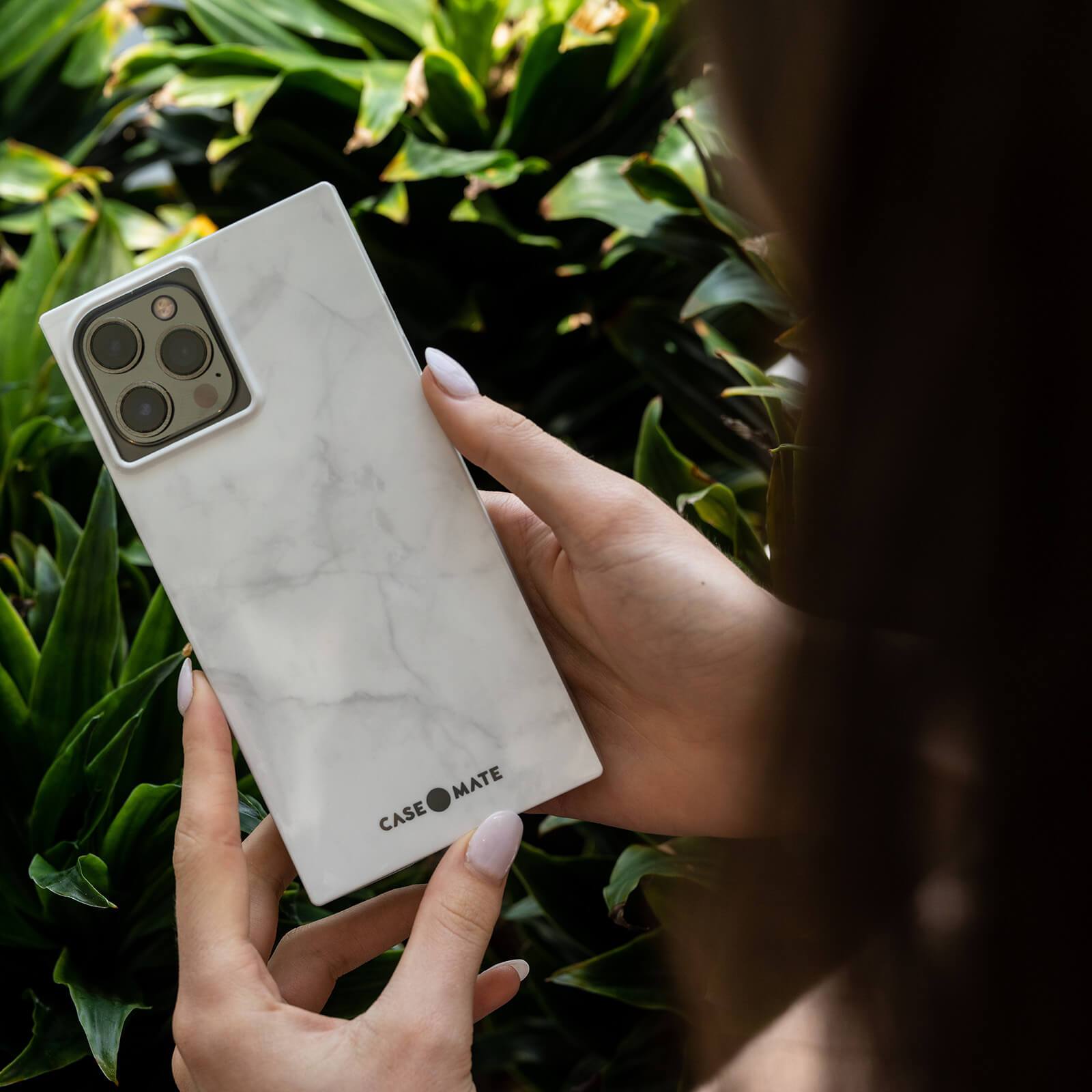 Hands holding White Marble case in front of a bush. color::White Marble
