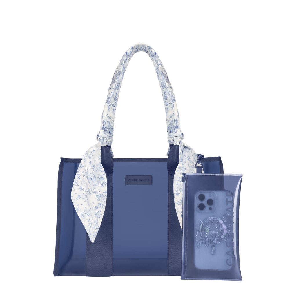 A navy blue tote bag with a white and blue floral patterned scarf tied to its handles, shown next to a matching translucent navy blue phone pouch on a solid white background. 
#color_something navy twilly 