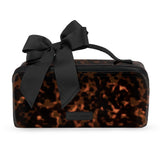 A rectangular cosmetic pouch with a black and brown tortoiseshell pattern, featuring a large black satin bow on top and a small black brand plaque on the front.
#color_tortoiseshell 