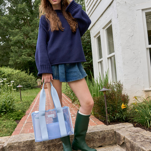 A woman stands on stone steps in a garden setting, wearing an oversized navy sweater, denim shorts, and green rain boots while holding a light blue translucent tote bag with white handles.
#color_carolina blue crochet wave