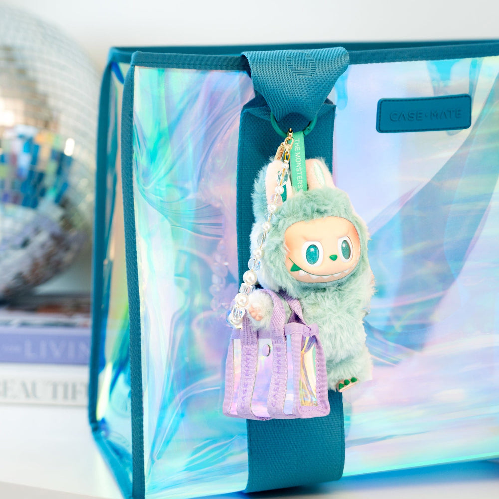 Plush keychain with a green creature design in front of a transparent box with a blue ribbon. #color_iridescent