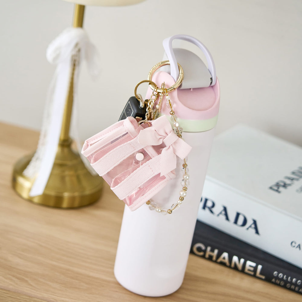 Pink shoe keychain with decorative elements on a white stand, with Prada and Chanel books in the background.
#color_ballet pink bow