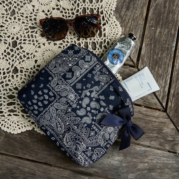 A navy blue and white paisley bandana zip pouch with a matching ribbon bow, styled on a rustic wooden table with a lace doily, sunglasses, a hand sanitizer bottle, and a small card.
#color_navy bandana bow