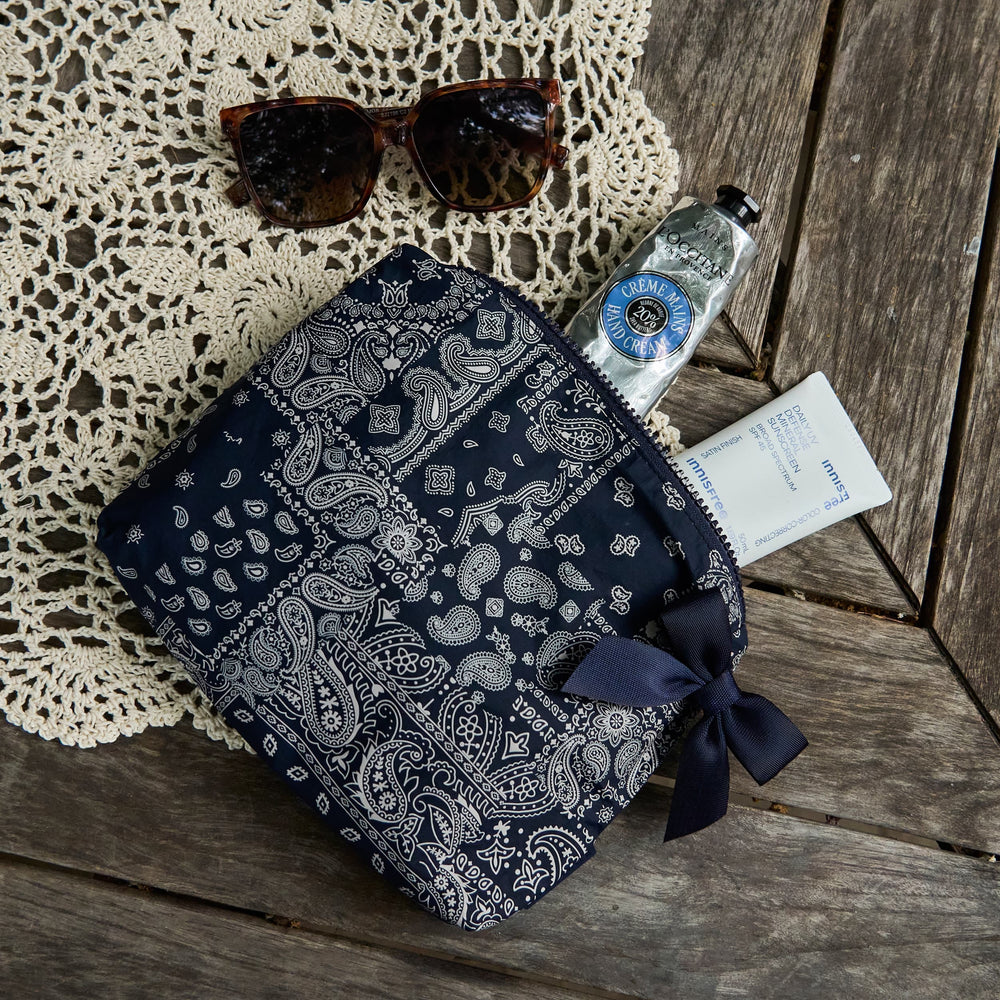 A navy blue and white paisley bandana zip pouch with a matching ribbon bow, styled on a rustic wooden table with a lace doily, sunglasses, a hand sanitizer bottle, and a small card.
#color_navy bandana bow
