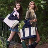 Two women standing outdoors in a wooded setting, each carrying a large tote bag with a wide ribbon handle. The tote bags are cream with navy blue panels and handles.
#color_oxford navy bow