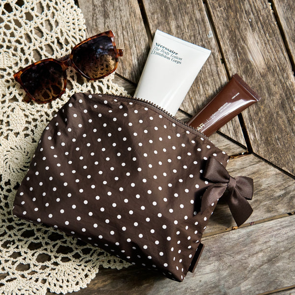 A brown cosmetic pouch with white polka dots and a matching bow, styled on a wooden surface with sunglasses, a tube of cream, and a brown tube.

#color_espresso polka dot bow