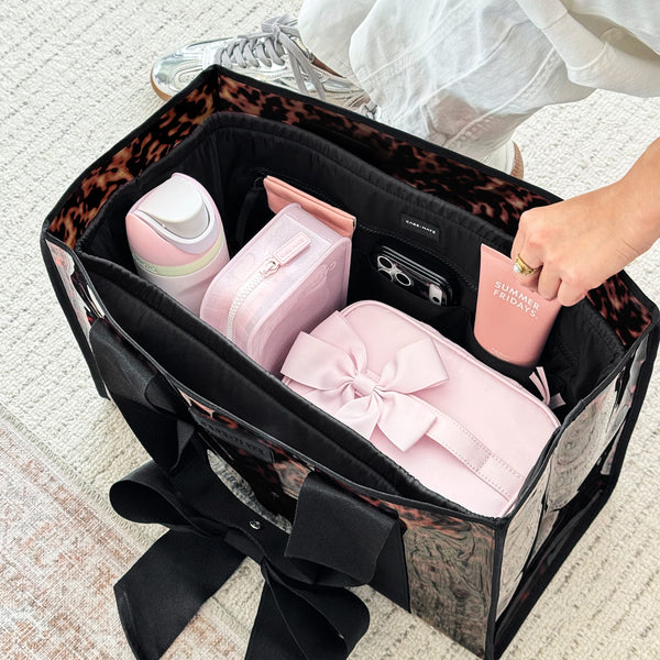 A black and leopard-print travel organizer tote bag filled with pink accessories, shown on a light textured floor with a person's hand placing a pink cup inside.

#color_midnight black