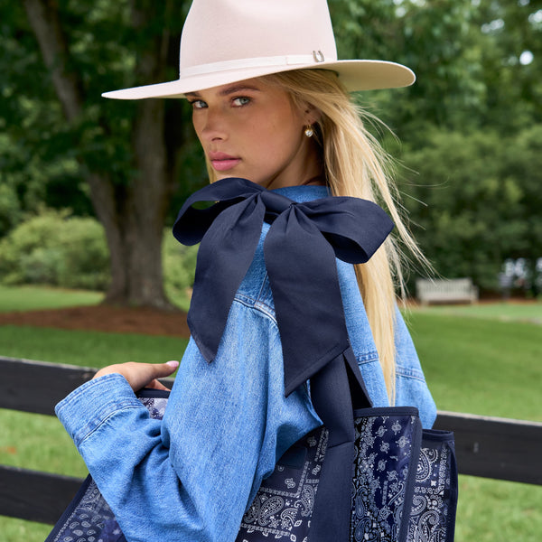 A woman outdoors wearing a wide-brim light beige hat, a light blue denim shirt, and carrying a navy blue bandana tote bag with a large navy bow.
#color_navy bandana bow