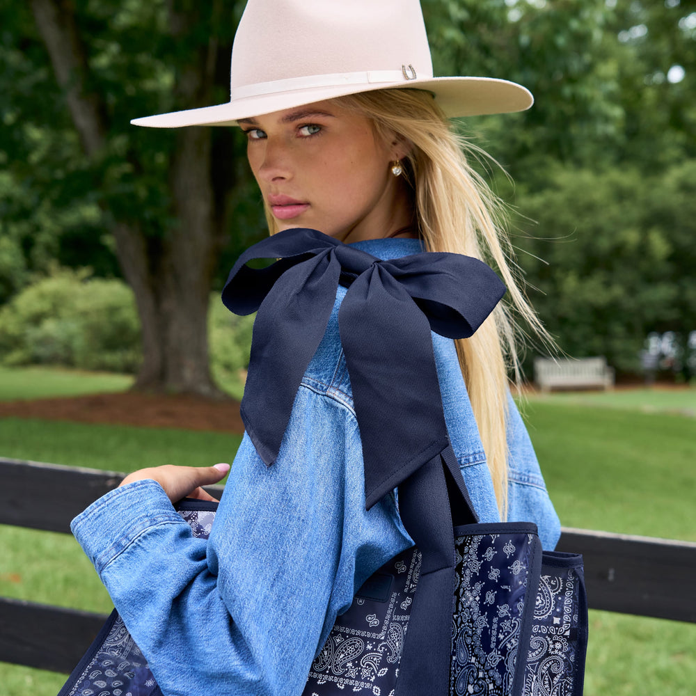 A woman outdoors wearing a wide-brim light beige hat, a light blue denim shirt, and carrying a navy blue bandana tote bag with a large navy bow.
#color_navy bandana bow