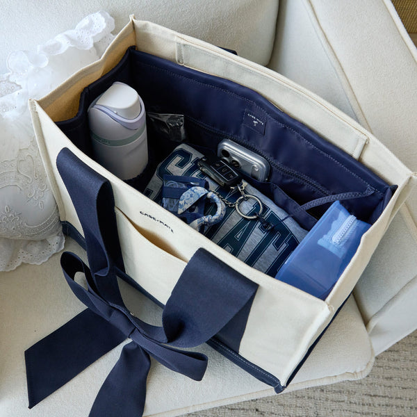 A cream canvas tote bag organizer with navy ribbon handles and interior pockets, shown filled with travel accessories on a light upholstered chair.
#color_navy