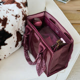 A translucent burgundy zippered organizer bag with patterned fabric handles, shown open on a white sofa next to a cow-print pillow.
#color_mulberry