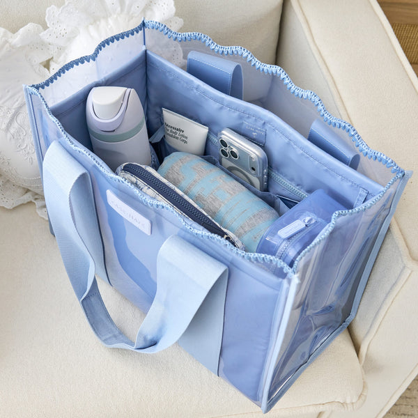 A light blue translucent tote bag with matching fabric handles, shown open and filled with toiletries and accessories on a cream-colored sofa.

#color_carolina blue