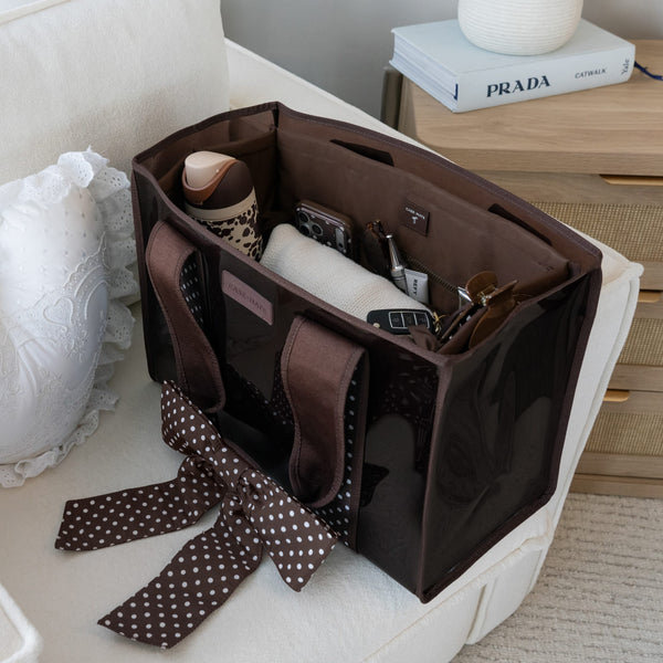 A brown tote organizer bag with a glossy finish and matching brown polka-dot ribbon handle, shown open on a white armchair with various accessories inside. A Prada book is visible on a wooden side table in the 
background.

#color_espresso