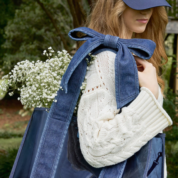 A woman outdoors wearing a large blue denim strap jelly tote bag with a wide shoulder strap tied in a bow over her shoulder, paired with a cream cable-knit sweater and a navy cap. White baby's breath flowers are tucked into the side of the bag.
#color_dark wash denim bow