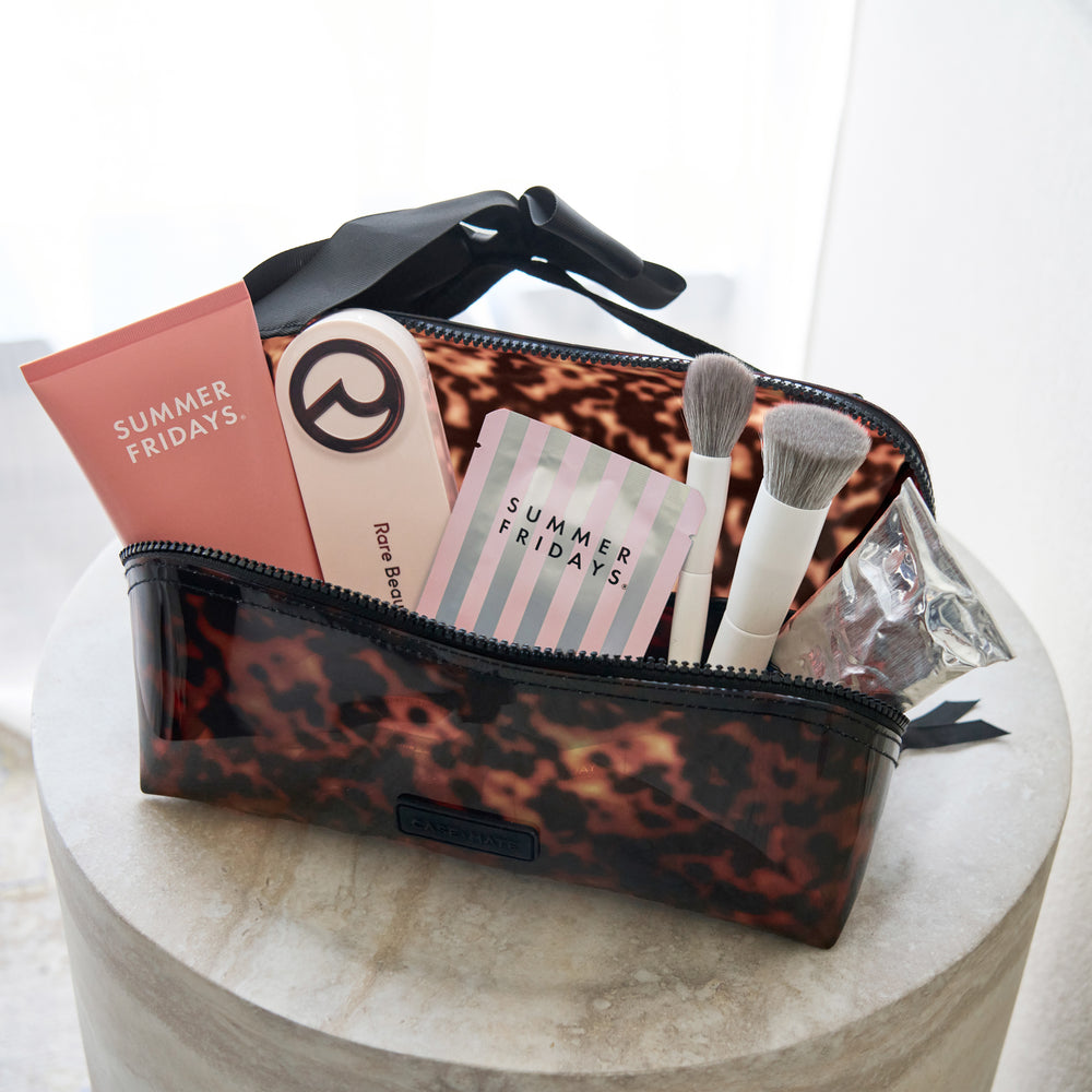 A clear cosmetic pouch with a brown leopard print pattern and black trim, shown open on a round stone pedestal. Inside are makeup brushes, a pink tube labeled 'SUMMER FRIDAYS,' a striped card, and a small clear bag.
#color_tortoiseshell 