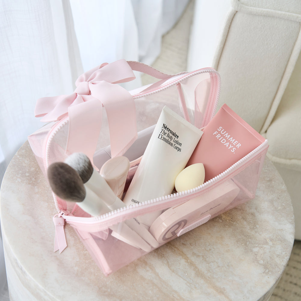 A pink translucent makeup bag with a large bow, filled with cosmetics, resting on a round marble table in a softly lit room.
#color_ballet pink bow