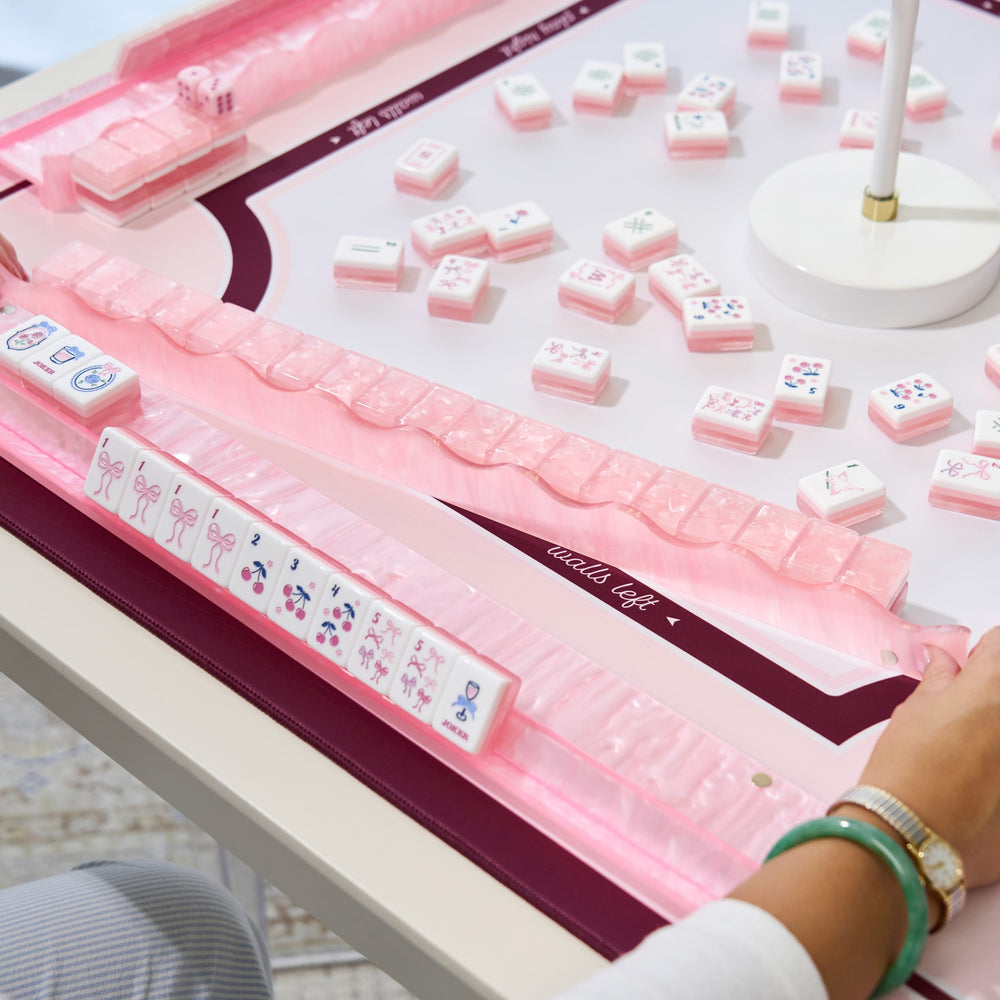 A close-up of a pink mahjong game set on a white table, with several tiles laid out and a person's hand visible at the lower right edge.
#color_Ballet Pink Pearl