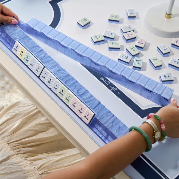 A close-up of a person's hands arranging a set of white and pastel-colored mahjong tiles on a blue mat over a white table. The person is wearing a beige dress and green and pink bracelets.
#color_carolina blue pearl