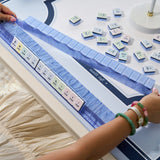A close-up of a person's hands arranging a set of white and pastel-colored mahjong tiles on a blue mat over a white table. The person is wearing a beige dress and green and pink bracelets.
#color_carolina blue pearl