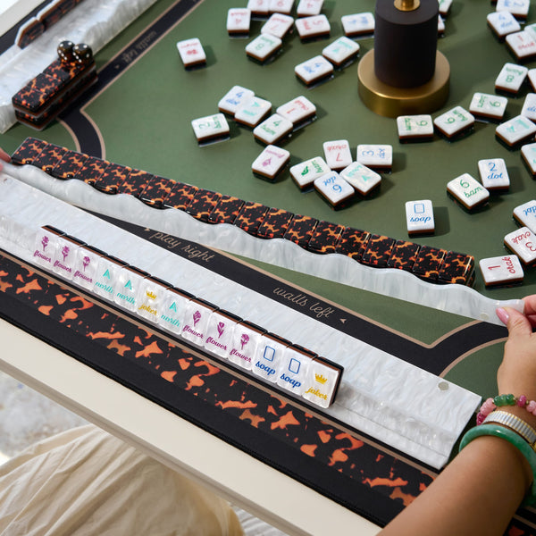 A close-up of a mahjong game table with tiles laid out, decorative black-and-orange patterned racks, dice, and a visible person's hand wearing green bracelets at the lower right.
#color_ivory pearl