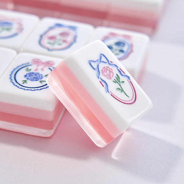 A close-up of a pink and white mahjong tile with a floral crest design, set against a matching pink and white background of other tiles.
#color_ballet pink pearl edition