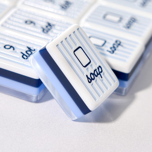 A close-up of a white and blue striped rectangular soap bar labeled 'soap' with a soap-bar icon, resting in front of several similar soap bars in their clear plastic packaging.
#color_cabana stripe edition